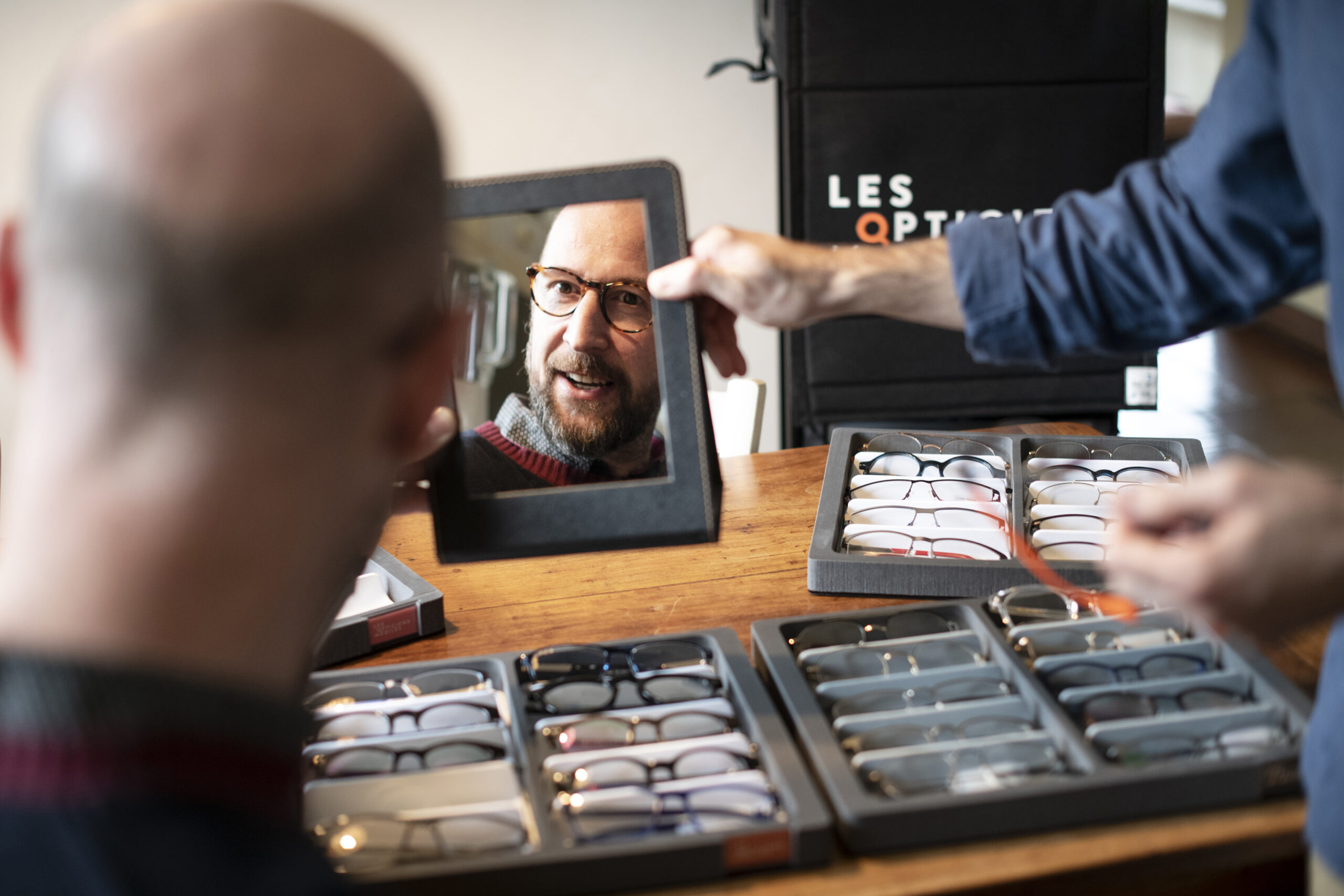 Client essayant de nouvelles montures de lunettes chez lui avec son opticien. Reflet souriant dans un miroir, entouré de montures de lunettes. Expérience personnalisée de sélection de lunettes avec l'aide d'un professionnel de santé Enseigne 'Les Opticiens Mobiles' visible en arrière-plan